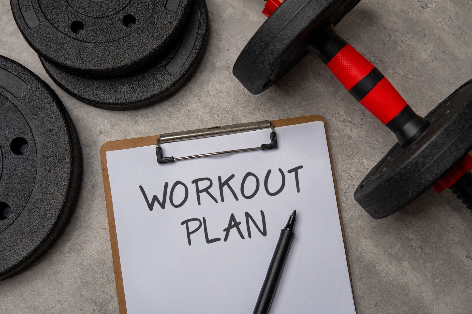 A close-up image of a clipboard with a WORKOUT PLAN written on it next to a black dumbbell and weight plates.