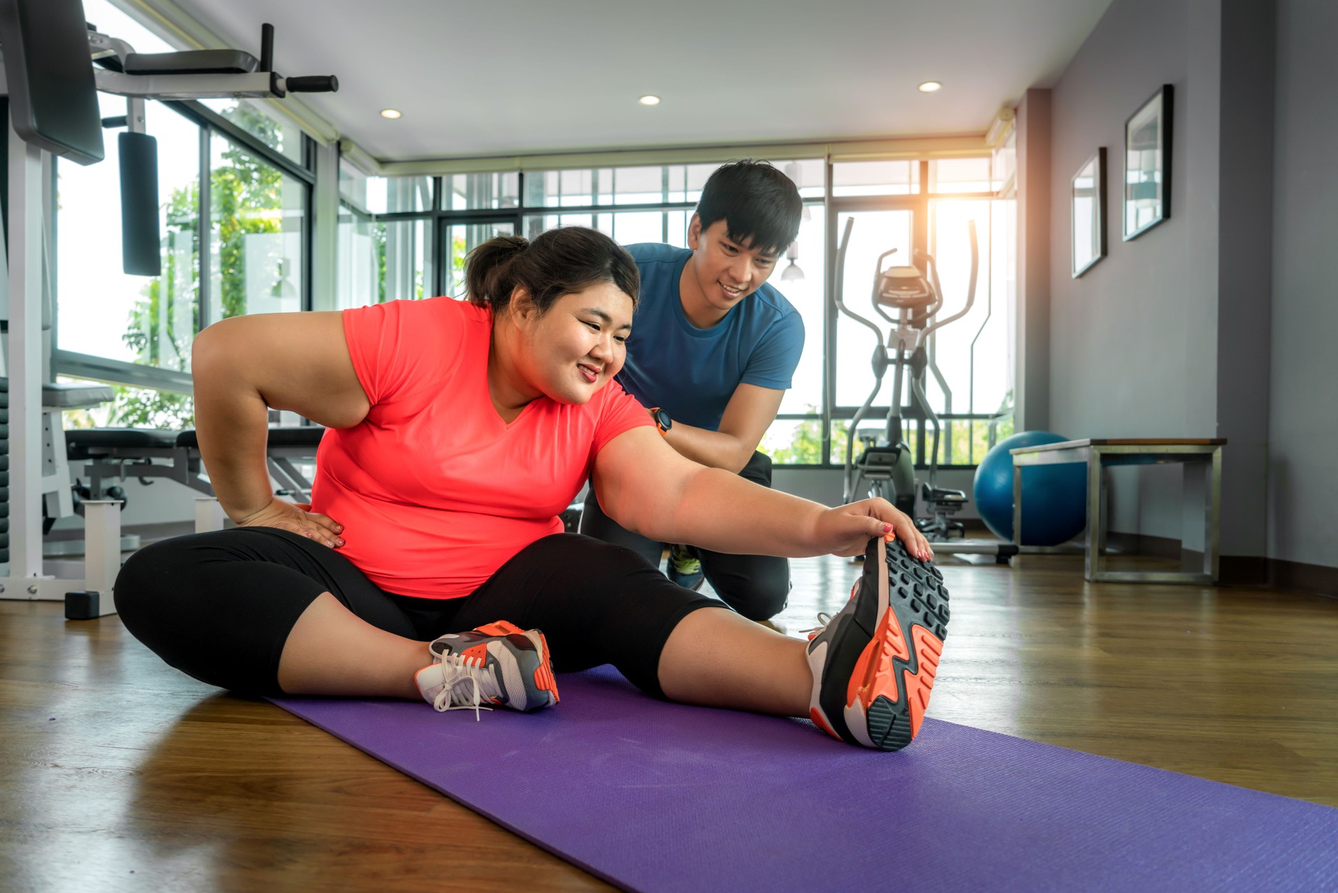 Two Asian trainer man and Overweight woman exercising stretch together in modern gym, happy and smile during workout. Fat women take care of health and want to lose weight concept.